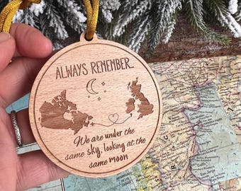 Könnte beinhalten: Ein runder Holzanhänger mit einer Karte von Kanada und Großbritannien. Der Anhänger hat ein goldenes Band und den Text "ALWAYS REMEMBER... We are under the same sky, looking at the same moon" ist darauf graviert.