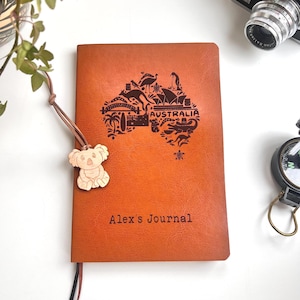 May include: A tan leather journal featuring a map of Australia etched on the cover, with the word "Australia" above the map. The journal is inscribed with "Alex's Journal". A wooden koala bear charm is attached to the spine.