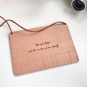 May include: A light brown wooden plaque with a leather cord hanging from the top. The plaque has the text "You are loved and the centre of our family" engraved on it.