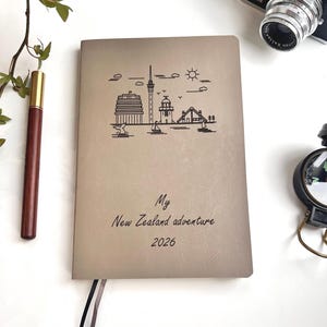 May include: Tan-colored journal with a black line drawing of a New Zealand cityscape. The text "My New Zealand adventure 2026" is printed below the drawing. A pen, camera, and compass are visible.