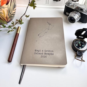 May include: A taupe-colored notebook with a map of New Zealand and the text "Magi a Gethin Seland Newydd 2026" printed on the cover. A pen, a vintage camera, and a compass are also visible.