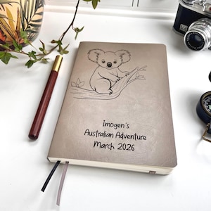 Könnte beinhalten: Beiges Notizbuch mit einer Koala-Illustration und dem Text "Imogen's Australian Adventure March 2026." Ein brauner Stift mit goldener Spitze liegt neben dem Notizbuch. Das Notizbuch hat zwei Lesebändchen.