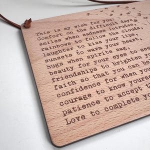 May include: A wooden plaque with a message about wishing comfort, smiles, laughter, sunsets, hugs, beauty, friendships, faith, confidence, courage, patience, and love. The plaque is hanging from a brown leather cord.