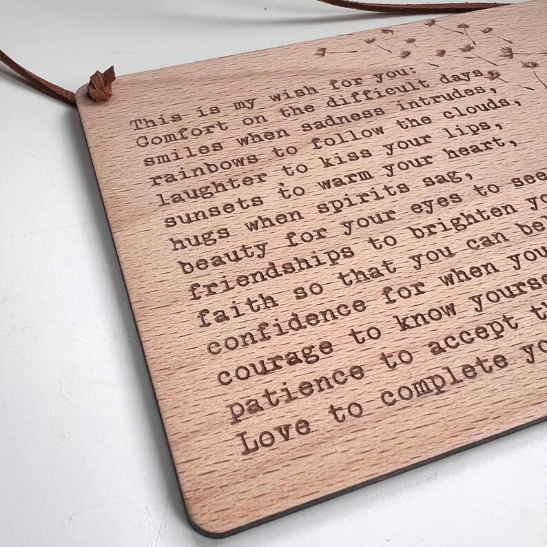 May include: A wooden plaque with a message about wishing comfort, smiles, laughter, sunsets, hugs, beauty, friendships, faith, confidence, courage, patience, and love. The plaque is hanging from a brown leather cord.