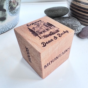 May include: A wooden cube with engraved text and a camper van design. The top of the cube reads "Exploring Since 2018" and "Dean & Becky." The side says "Mountains" and "Woods."