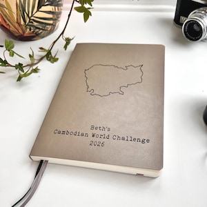 Puede incluir: Un cuaderno beige con el contorno de Camboya en la portada. El texto "Beth's Cambodian World Challenge 2026" está impreso debajo del mapa. El cuaderno tiene una cinta de marcador.
