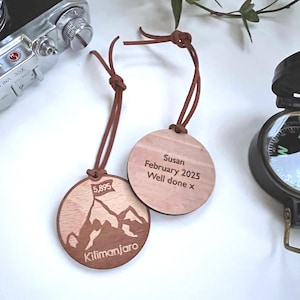 May include: Two wooden round ornaments with brown leather cords. One ornament features a mountain silhouette with the text "5,895" and "Kilimanjaro". The other ornament has the text "Susan February 2025 Well done x".