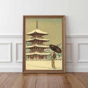 May include: Framed art print depicting a Japanese pagoda in a snowy landscape. The artwork features a multi-tiered pagoda with a person holding an umbrella. The color palette includes shades of brown, beige, and white, creating a serene and traditional aesthetic.