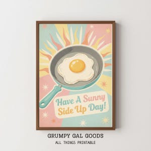 May include: A framed print featuring a retro-style illustration of a fried egg in a pan. The artwork has a pastel color scheme with the text "Have A Sunny Side Up Day!" and the brand name "Grumpy Gal Goods".