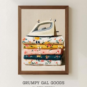 May include: A framed print featuring a stack of folded floral fabric with a vintage iron on top. The fabric is in shades of white, yellow, pink, and teal. The print has the text "GRUMPY GAL GOODS ALL THINGS PRINTABLE".