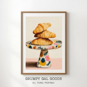 May include: A framed print featuring three golden-brown croissants stacked on a decorative cake stand. The stand has a floral and geometric pattern. The print is labeled "GRUMPY GAL GOODS ALL THINGS PRINTABLE."