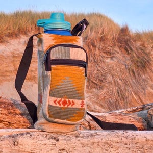 May include: A water bottle with a teal lid and a patterned fabric carrier. The carrier has a small zippered pouch and a black shoulder strap. The fabric design features orange, green, and red geometric patterns. The bottle is blue.