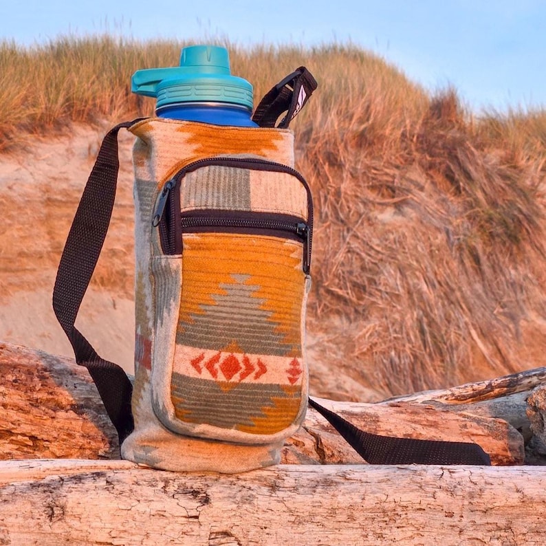 May include: A water bottle with a teal lid and a patterned fabric carrier. The carrier has a small zippered pouch and a black shoulder strap. The fabric design features orange, green, and red geometric patterns. The bottle is blue.