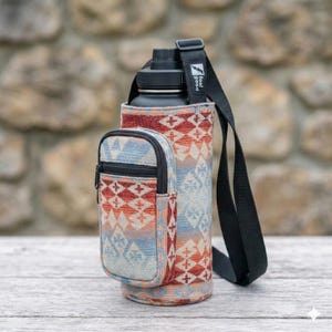 May include: A black water bottle in a patterned fabric carrier with a small zippered pouch. The carrier has a black shoulder strap and a design in shades of red, blue, and orange. The text "feel good" is on the strap.