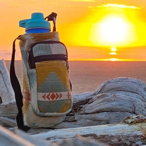 May include: A water bottle with a teal lid and a patterned fabric carrier. The carrier has a small zipped pocket and a black shoulder strap. The background shows a sunset over the sea.