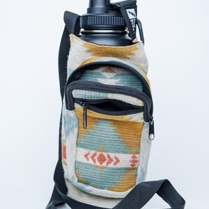 May include: A black water bottle in a patterned fabric carrier with a shoulder strap. The carrier has a zippered pocket and a design in shades of blue, orange, and yellow. The bottle has a black cap.