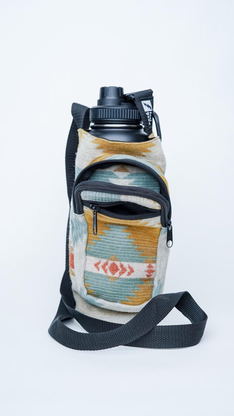 May include: A black water bottle in a patterned fabric carrier with a shoulder strap. The carrier has a zippered pocket and a design in shades of blue, orange, and yellow. The bottle has a black cap.