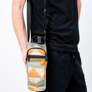 May include: A water bottle carrier with a southwestern pattern in shades of green, yellow, and beige. The carrier has a small zipped pocket and a black adjustable strap. The bottle is black.