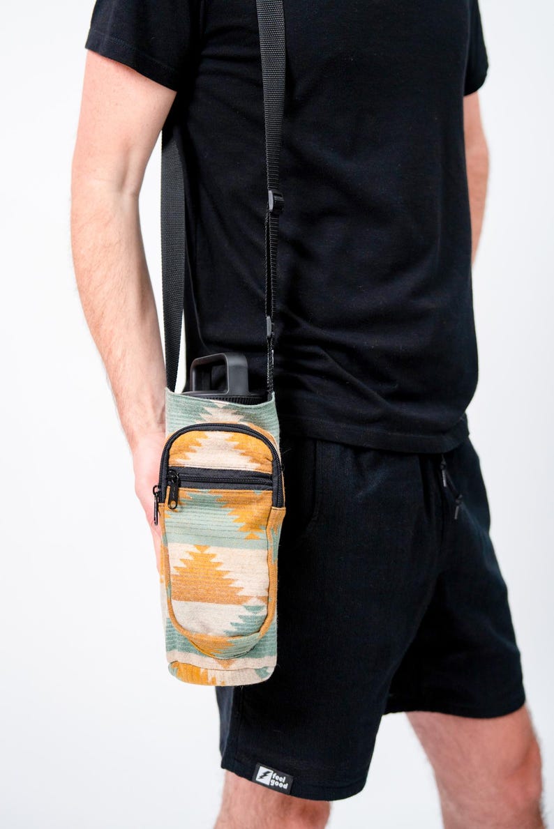 May include: A water bottle carrier with a southwestern pattern in shades of green, yellow, and beige. The carrier has a small zipped pocket and a black adjustable strap. The bottle is black.