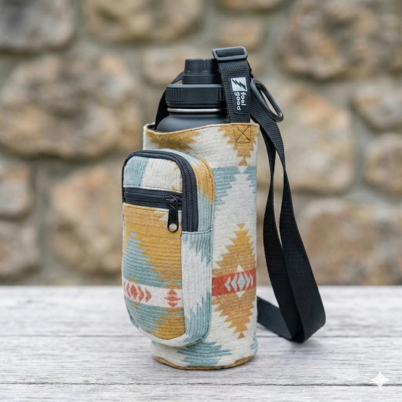May include: A black water bottle in a patterned fabric carrier with a small zipped pocket. The carrier features a geometric design in shades of gold, blue, and red. A black strap is attached to the carrier. The text "feel good" is on the strap.