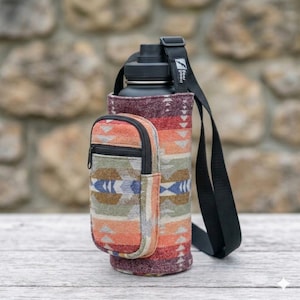 May include: A black water bottle in a patterned fabric carrier with a small zippered pouch. The carrier has a black shoulder strap and is decorated with a Southwestern-inspired design in shades of orange, red, and blue.