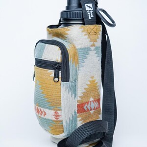 May include: Water bottle holder with a southwestern design in yellow, blue, and red. It features a small zipped pocket and a black shoulder strap. The text "feel good" is printed on a black tag.