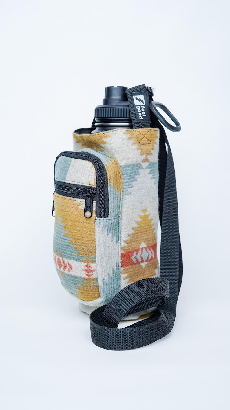 May include: Water bottle holder with a southwestern design in yellow, blue, and red. It features a small zipped pocket and a black shoulder strap. The text "feel good" is printed on a black tag.