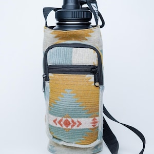 May include: A black water bottle in a patterned sleeve with a zip pocket and a black shoulder strap. The sleeve has a geometric design in yellow, blue, and red.