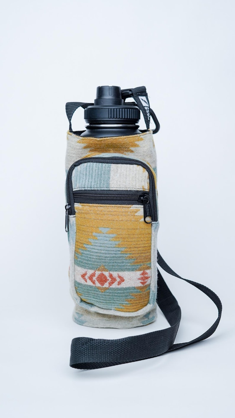 May include: A black water bottle in a patterned sleeve with a zip pocket and a black shoulder strap. The sleeve has a geometric design in yellow, blue, and red.