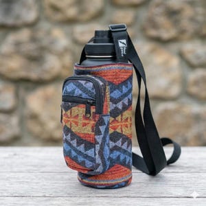 May include: Black water bottle with a patterned fabric sleeve in shades of red, blue, and gold. The sleeve has a zippered pocket and a black shoulder strap. The text "feel good" is printed on the strap.