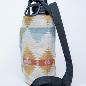 May include: A water bottle with a black lid and strap, encased in a patterned fabric sleeve. The sleeve features a geometric design in yellow, orange, and blue on a neutral background. The black strap is adjustable.