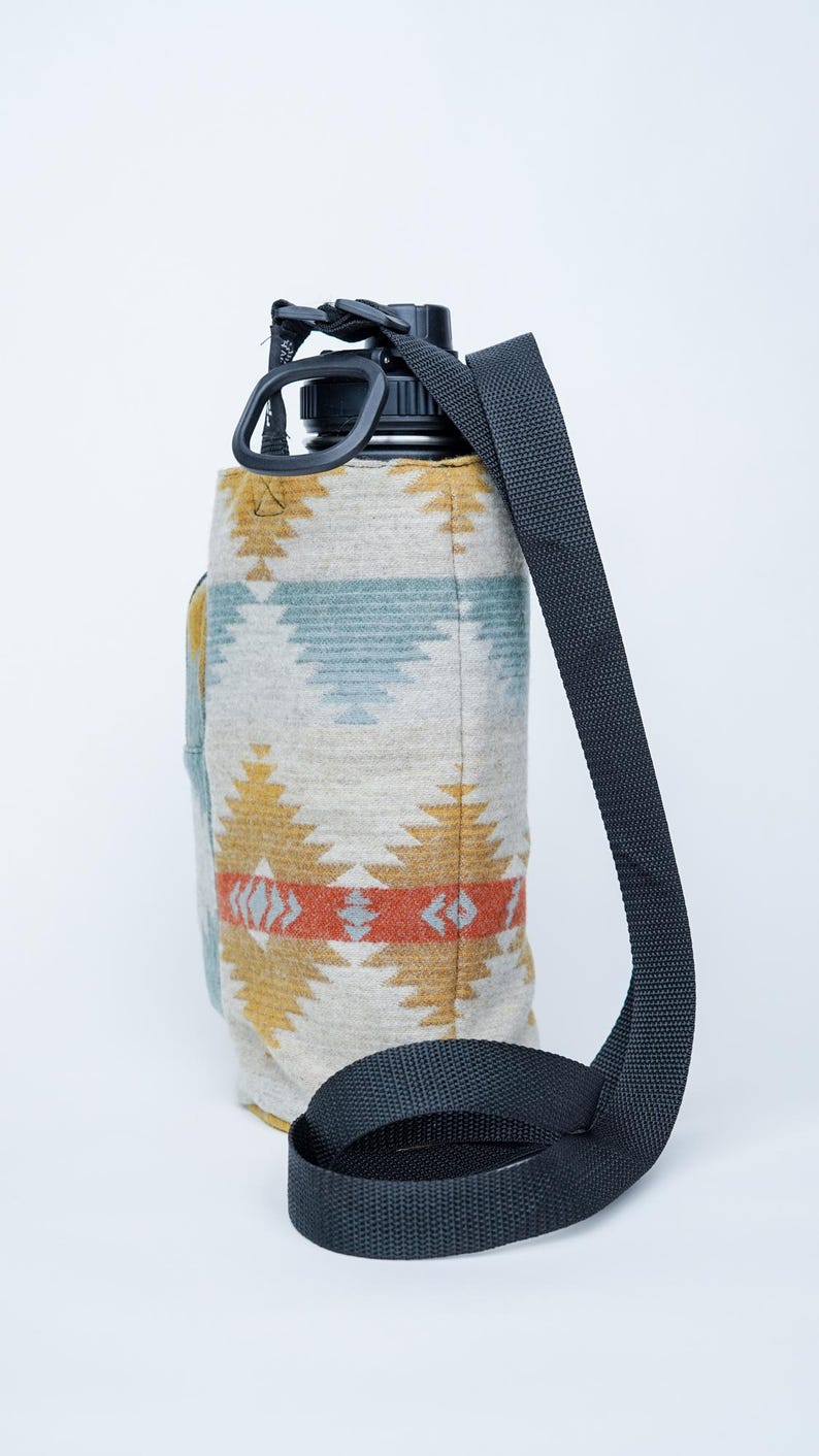 May include: A water bottle with a black lid and strap, encased in a patterned fabric sleeve. The sleeve features a geometric design in yellow, orange, and blue on a neutral background. The black strap is adjustable.