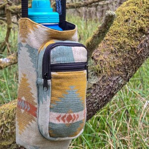 May include: A water bottle holder with a southwestern design in beige, yellow, teal, and orange. It features a small zipped pocket and a black strap. The water bottle has a blue lid. The holder is suspended from a tree branch.