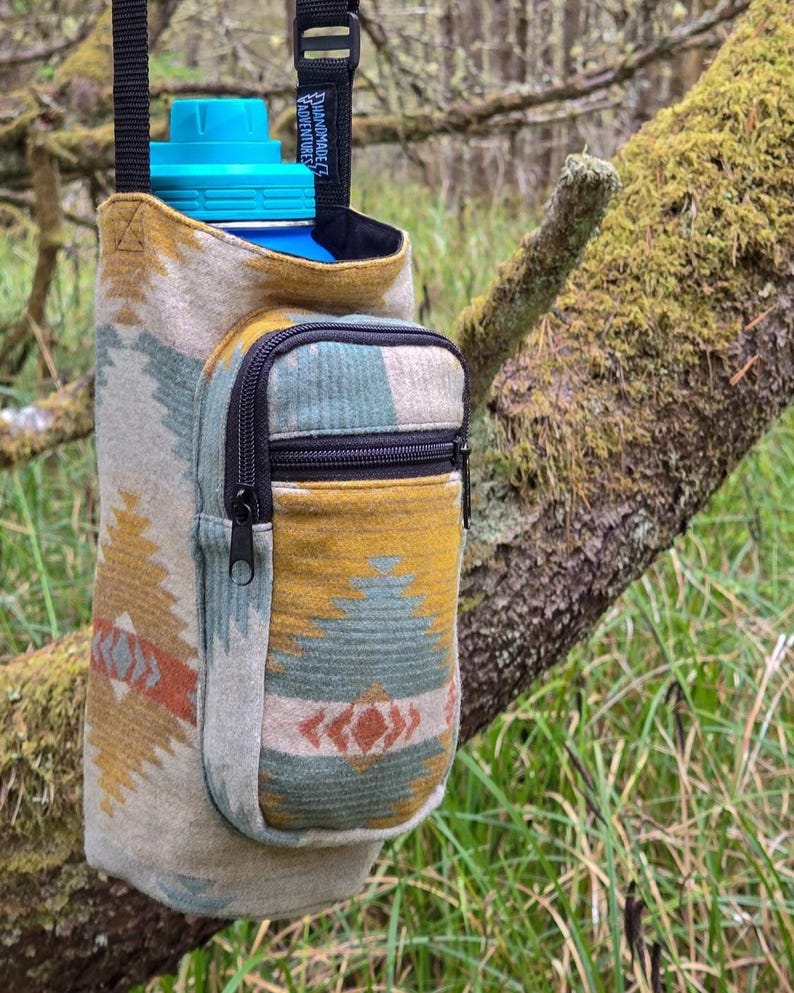 May include: A water bottle holder with a southwestern design in beige, yellow, teal, and orange. It features a small zipped pocket and a black strap. The water bottle has a blue lid. The holder is suspended from a tree branch.