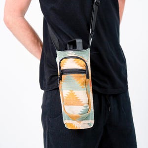 May include: A water bottle carrier with a southwestern pattern in teal, orange, and cream. It features a zippered pocket and a black adjustable shoulder strap. The bottle is black.