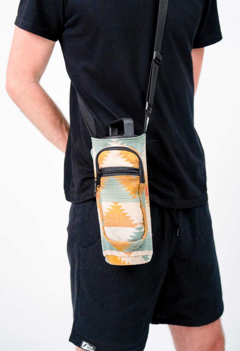 May include: A water bottle carrier with a southwestern pattern in teal, orange, and cream. It features a zippered pocket and a black adjustable shoulder strap. The bottle is black.