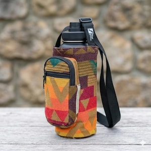 May include: A black water bottle with a black strap and a colorful patterned sleeve. The sleeve has a small zippered pouch and is decorated with geometric shapes in shades of orange, red, and brown. The text "Feel Good" is visible on the strap.