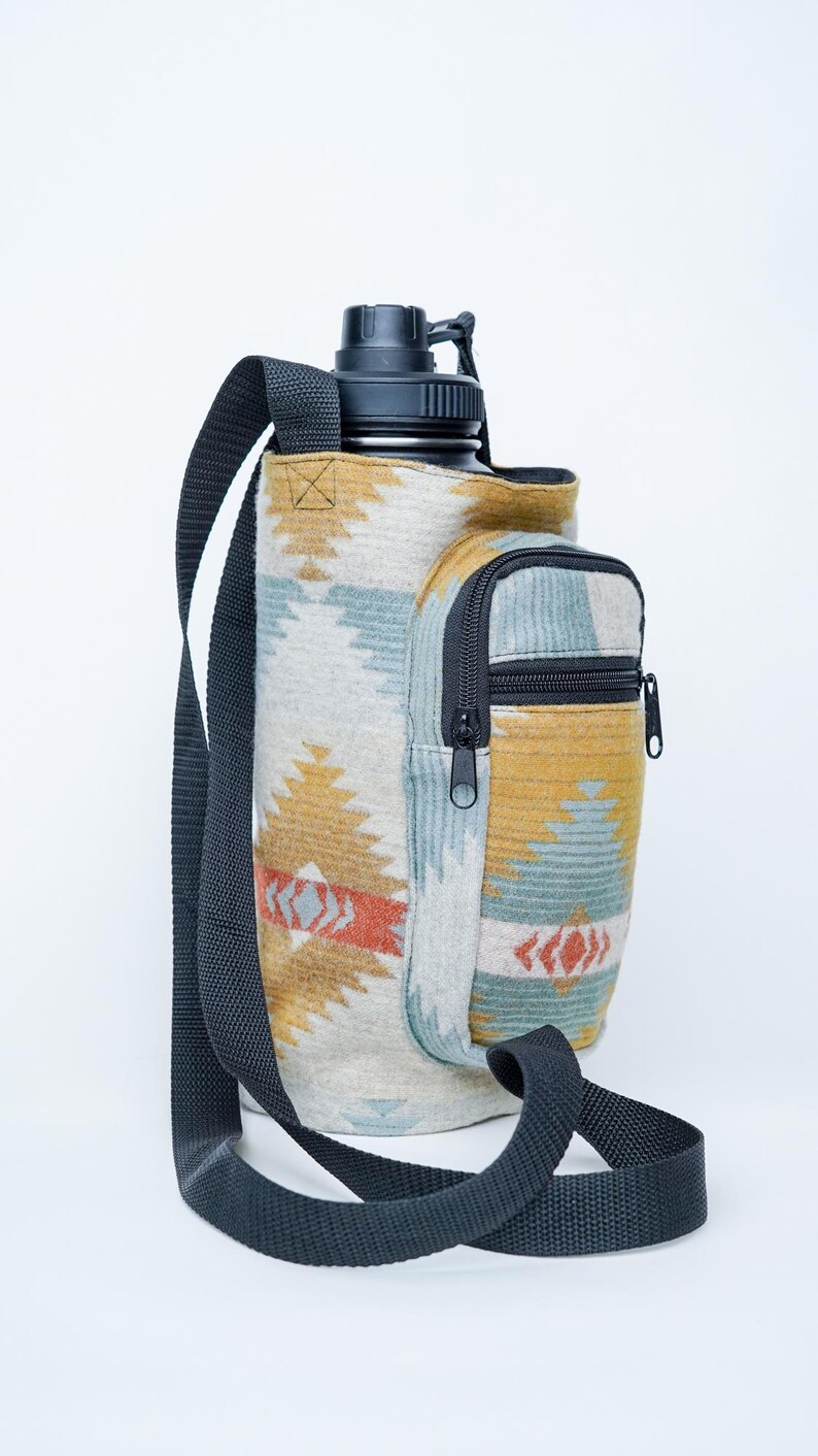 May include: A water bottle carrier with a black strap and a small zipped pocket. The carrier features a geometric pattern in beige, blue, orange, and yellow. The water bottle has a black lid.