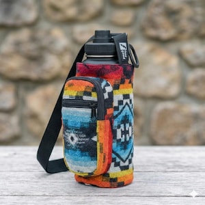 May include: A black water bottle with a colourful, patterned sleeve and a small zipped pouch. The sleeve features a geometric design in shades of blue, orange, red, and yellow. A black strap is attached for carrying. The text "feel good" is visible.