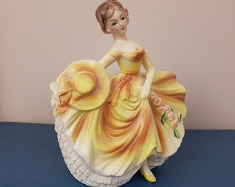 May include: A ceramic figurine of a woman in a yellow and orange dress with a large straw hat. She is holding a bouquet of flowers in her hand.