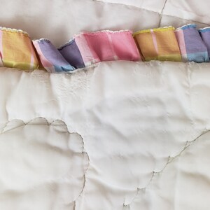 May include: A white quilted blanket with a pastel pink, yellow, and blue plaid trim. The blanket has a cloud-like quilted pattern.
