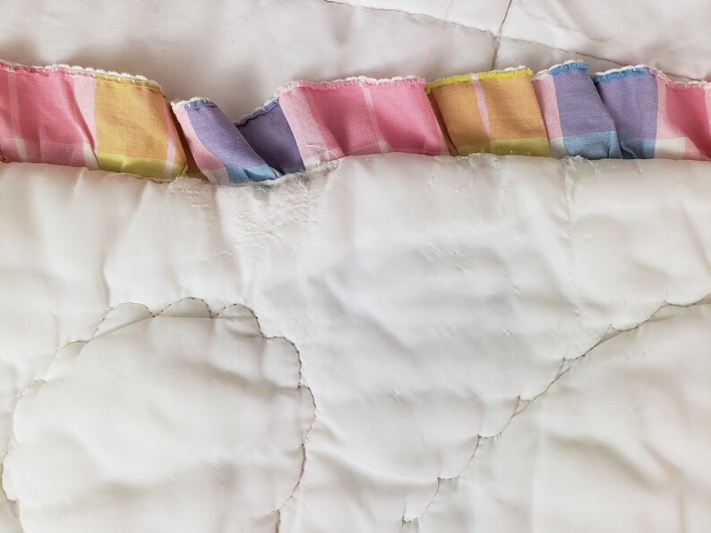 May include: A white quilted blanket with a pastel pink, yellow, and blue plaid trim. The blanket has a cloud-like quilted pattern.