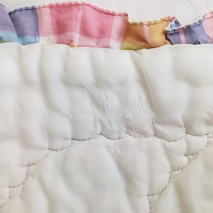 May include: White quilted fabric with a pink, yellow, and purple plaid trim. The fabric has a quilted pattern with stitching in a curved design.
