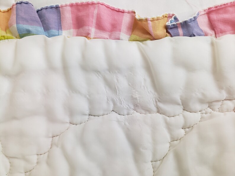 May include: White quilted fabric with a pink, yellow, and purple plaid trim. The fabric has a quilted pattern with stitching in a curved design.