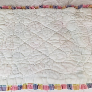 May include: White quilted baby blanket with a pink, yellow, blue, and purple plaid ruffle border.