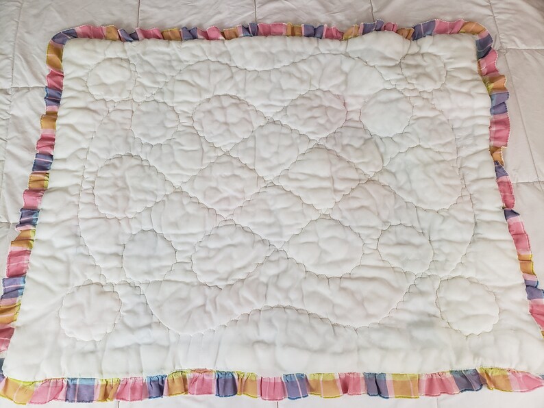 May include: White quilted baby blanket with a pink, yellow, blue, and purple plaid ruffle border.