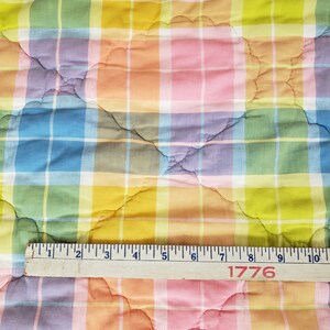 May include: A colorful plaid fabric with a quilted pattern. The fabric is in shades of pink, blue, yellow, and green. A wooden ruler with the number 1776 printed on it is placed on top of the fabric.