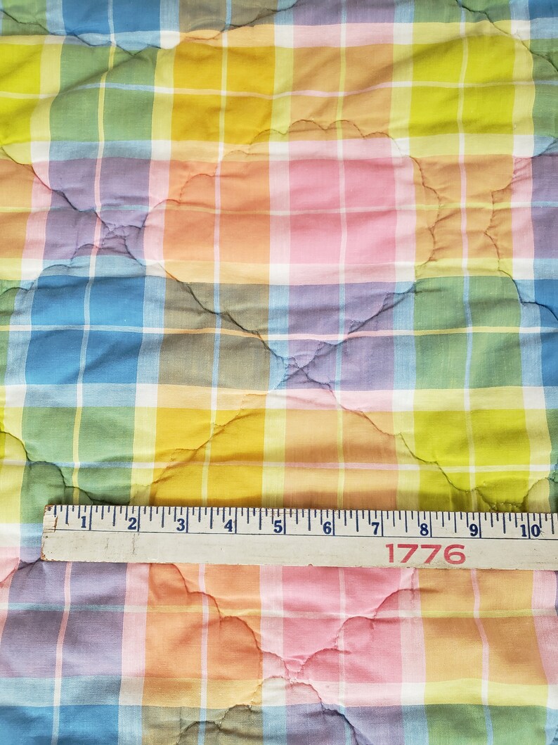 May include: A colorful plaid fabric with a quilted pattern. The fabric is in shades of pink, blue, yellow, and green. A wooden ruler with the number 1776 printed on it is placed on top of the fabric.