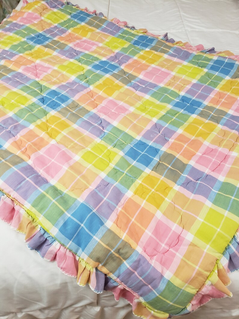 May include: A colorful plaid quilt with a ruffled edge. The quilt is made of a soft, cotton fabric and features a variety of pastel colors, including pink, blue, yellow, and green.