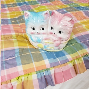 May include: Two ceramic kittens in a bathtub, one blue and one pink, on a multi-colored plaid fabric with a ruffled edge.